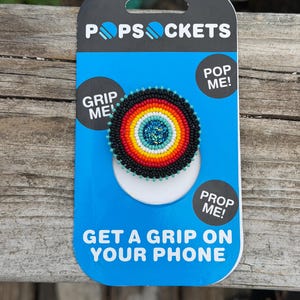 May include: A circular phone grip featuring concentric rings of black, red, orange, yellow, and white beads surrounding a central blue glitter disc. The product is mounted on a blue and black cardboard display card with the text POPSOCKETS.