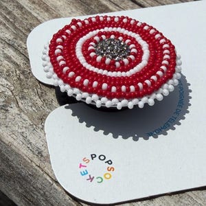 May include: A circular phone grip accessory featuring concentric rings of red and white seed beads with a sparkling silver center. The item is mounted on a white display card with the colorful text POPSOCKETS printed near the bottom edge.