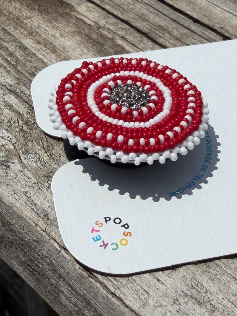 May include: A circular phone grip accessory featuring concentric rings of red and white seed beads with a sparkling silver center. The item is mounted on a white display card with the colorful text POPSOCKETS printed near the bottom edge.
