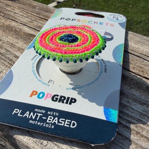 May include: A circular phone grip featuring a beaded top with concentric rings of neon pink, lime green, and a dark blue center. The grip is mounted on its original white cardboard packaging labeled PopSockets PopGrip made with plant-based materials.