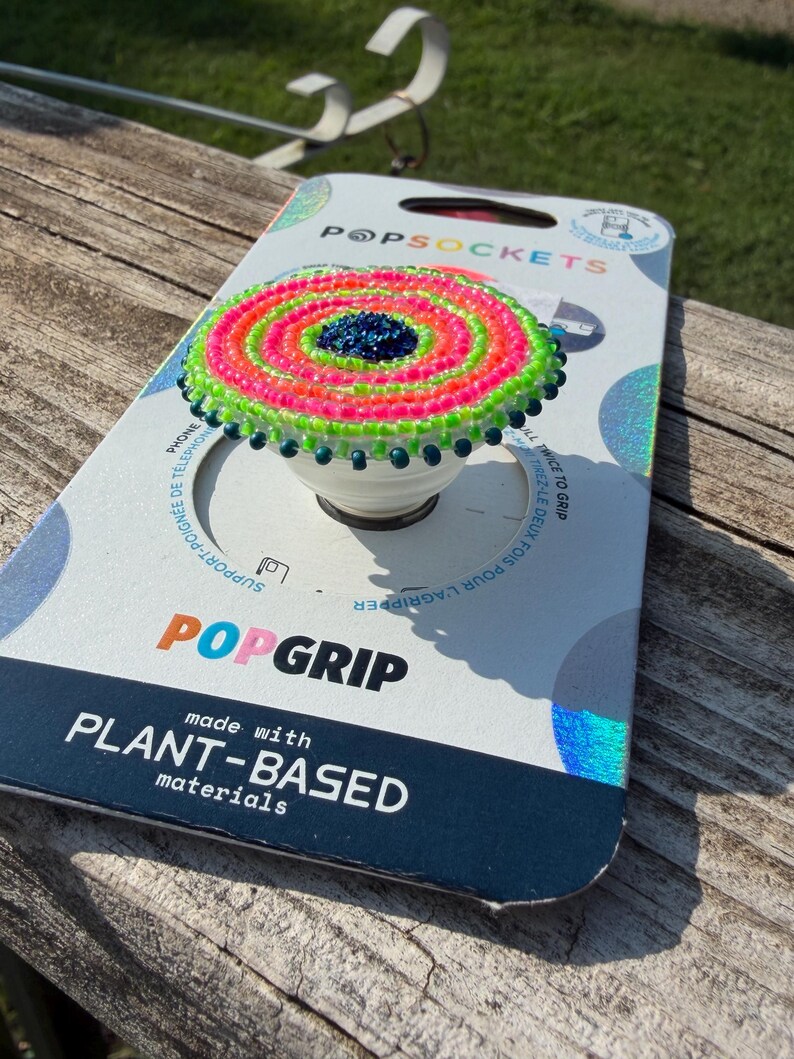 May include: A circular phone grip featuring a beaded top with concentric rings of neon pink, lime green, and a dark blue center. The grip is mounted on its original white cardboard packaging labeled PopSockets PopGrip made with plant-based materials.
