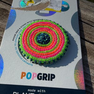 May include: A circular phone grip featuring concentric rings of neon green and bright pink seed beads surrounding a dark blue glitter center. The accessory is mounted on a white and holographic retail card labeled PopSockets and PopGrip.