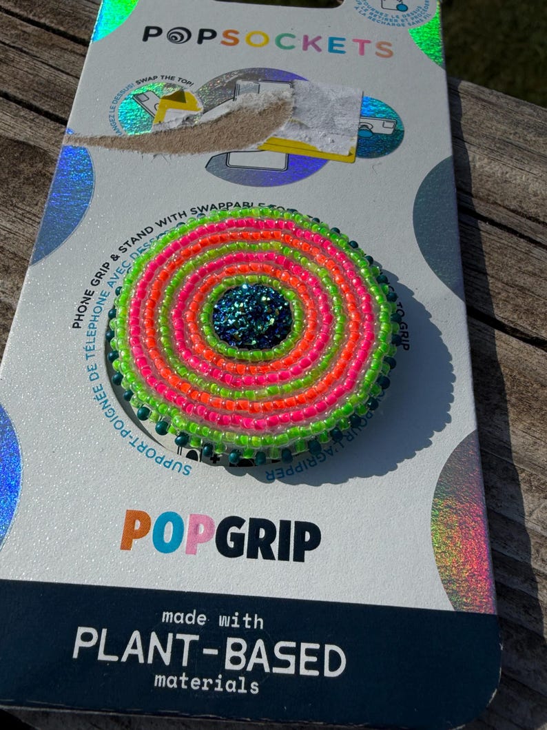 May include: A circular phone grip featuring concentric rings of neon green and bright pink seed beads surrounding a dark blue glitter center. The accessory is mounted on a white and holographic retail card labeled PopSockets and PopGrip.