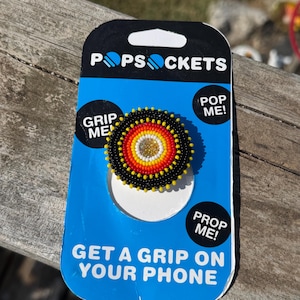 May include: A circular phone grip featuring a beaded design with concentric rings of black, red, orange, yellow, and a gold center. The grip is mounted on a blue and black PopSockets retail card with the text PopSockets, Grip Me, Pop Me, Prop Me, and Get A Grip On Your Phone.