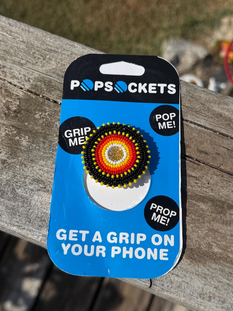 May include: A circular phone grip featuring a beaded design with concentric rings of black, red, orange, yellow, and a gold center. The grip is mounted on a blue and black PopSockets retail card with the text PopSockets, Grip Me, Pop Me, Prop Me, and Get A Grip On Your Phone.