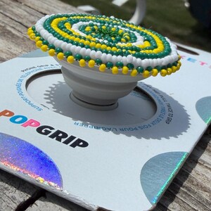 May include: A white phone grip featuring a circular top decorated with a spiral pattern of small yellow, green, and white beads. The device is mounted on a white cardboard backing with the text POPGRIP printed in black and blue lettering.
