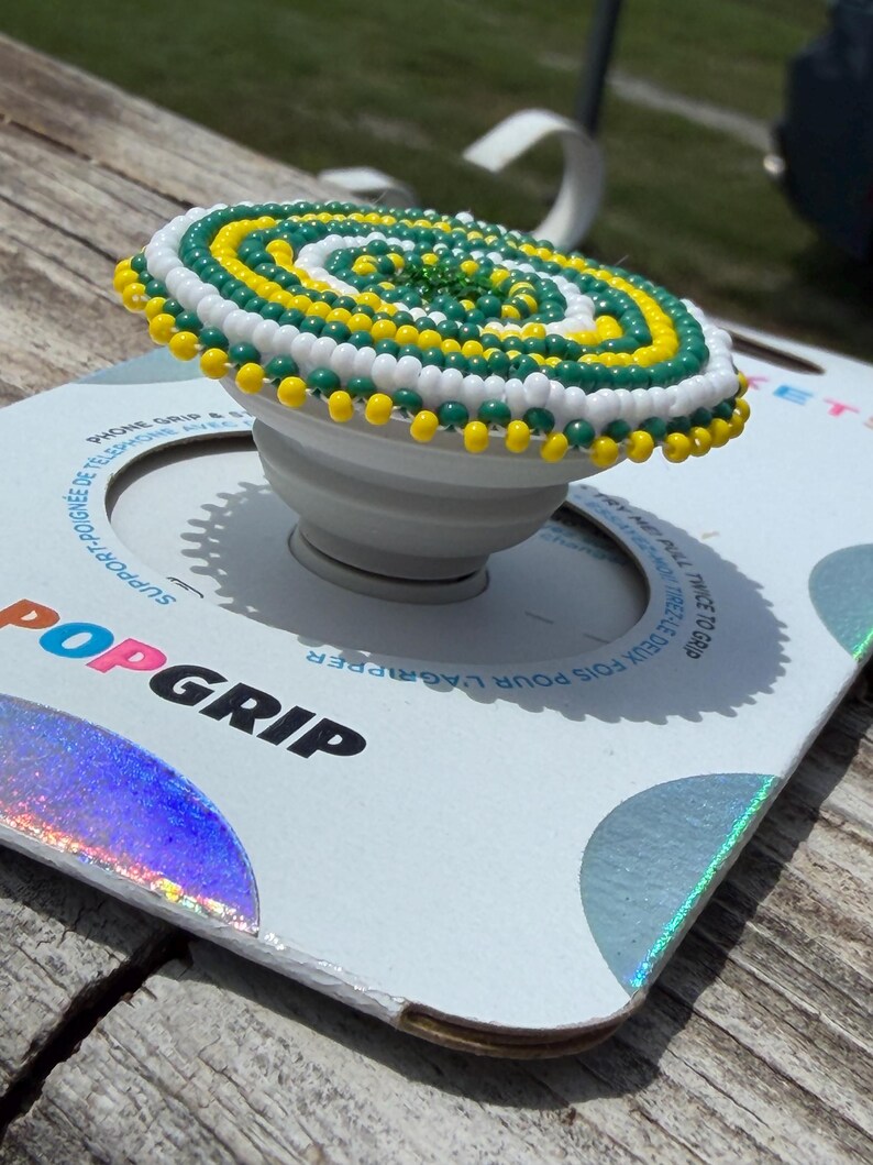 May include: A white phone grip featuring a circular top decorated with a spiral pattern of small yellow, green, and white beads. The device is mounted on a white cardboard backing with the text POPGRIP printed in black and blue lettering.