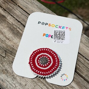 May include: A circular phone grip featuring a beaded design with concentric rings of red and white beads surrounding a central cluster of dark grey, shimmering beads. The grip is attached to a white rectangular display card with the text POPSOCKETS.