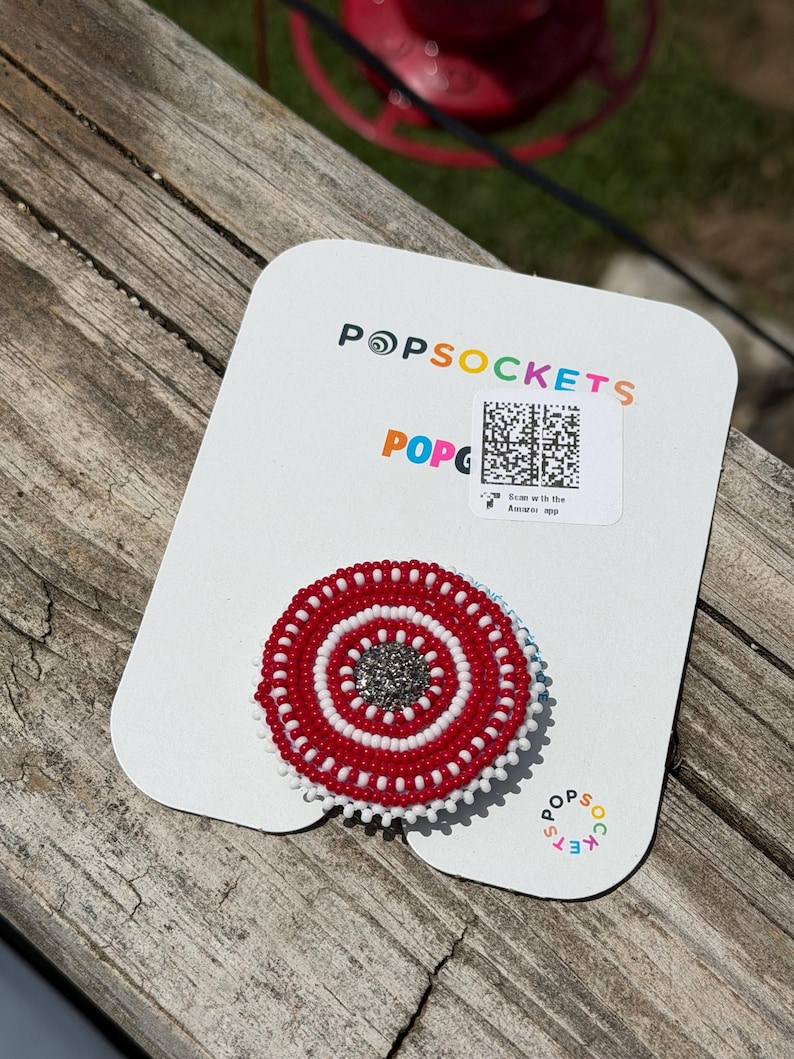 May include: A circular phone grip featuring a beaded design with concentric rings of red and white beads surrounding a central cluster of dark grey, shimmering beads. The grip is attached to a white rectangular display card with the text POPSOCKETS.