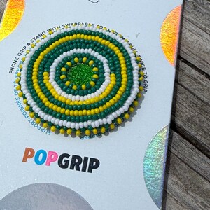 May include: A circular phone grip featuring a concentric pattern of small green, yellow, and white beads. The center is a solid bright green bead. The grip is mounted on a white display card with the text POPGRIP printed in black and orange.