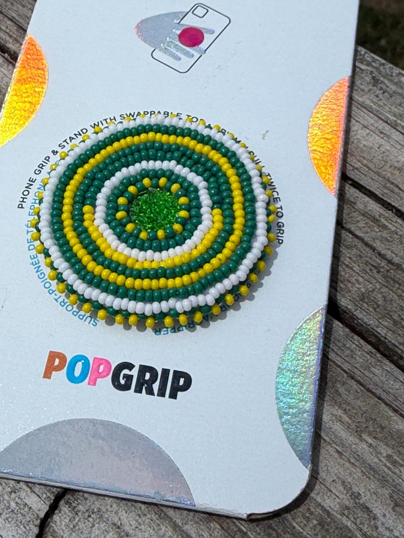 May include: A circular phone grip featuring a concentric pattern of small green, yellow, and white beads. The center is a solid bright green bead. The grip is mounted on a white display card with the text POPGRIP printed in black and orange.