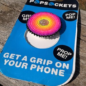 May include: A circular phone grip featuring a beaded design with a central silver glitter circle surrounded by concentric rings of yellow, orange, and pink beads, finished with a black beaded edge, mounted on blue and black PopSockets packaging.