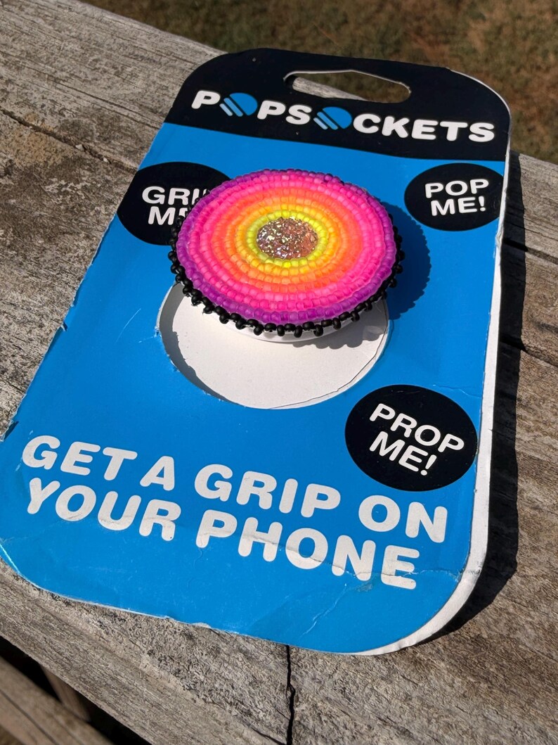 May include: A circular phone grip featuring a beaded design with a central silver glitter circle surrounded by concentric rings of yellow, orange, and pink beads, finished with a black beaded edge, mounted on blue and black PopSockets packaging.