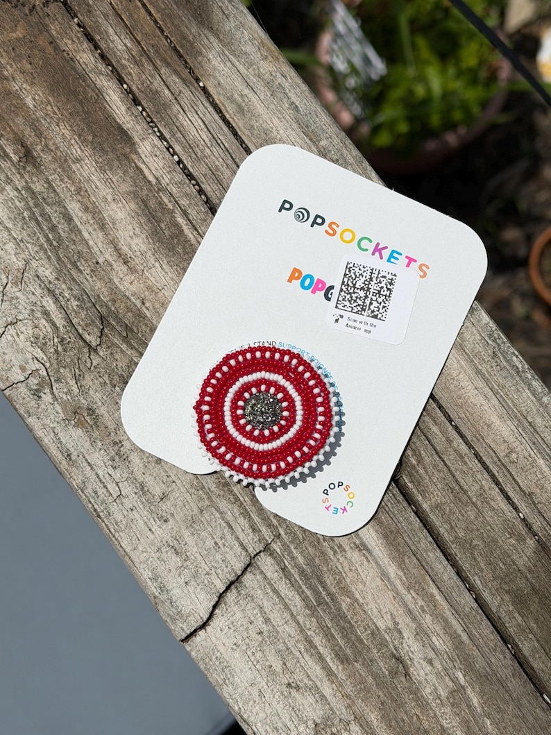 May include: A circular phone grip featuring a beaded design with concentric rings of red and white seed beads surrounding a dark gray center. The grip is attached to a white branded Popsockets display card with a QR code and colorful text.