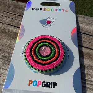 May include: A circular phone grip featuring concentric rings of neon pink, bright orange, lime green, and black seed beads surrounding a textured pink center. The accessory is mounted on a white PopSockets brand display card with Star Wars branding.