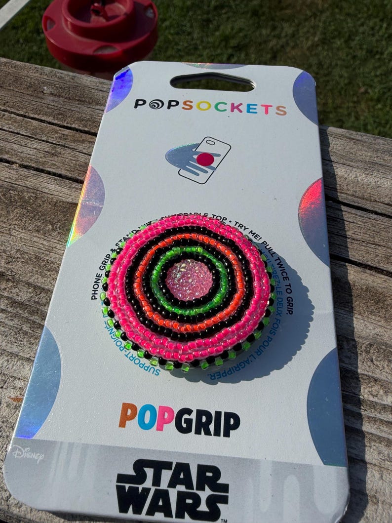 May include: A circular phone grip featuring concentric rings of neon pink, bright orange, lime green, and black seed beads surrounding a textured pink center. The accessory is mounted on a white PopSockets brand display card with Star Wars branding.