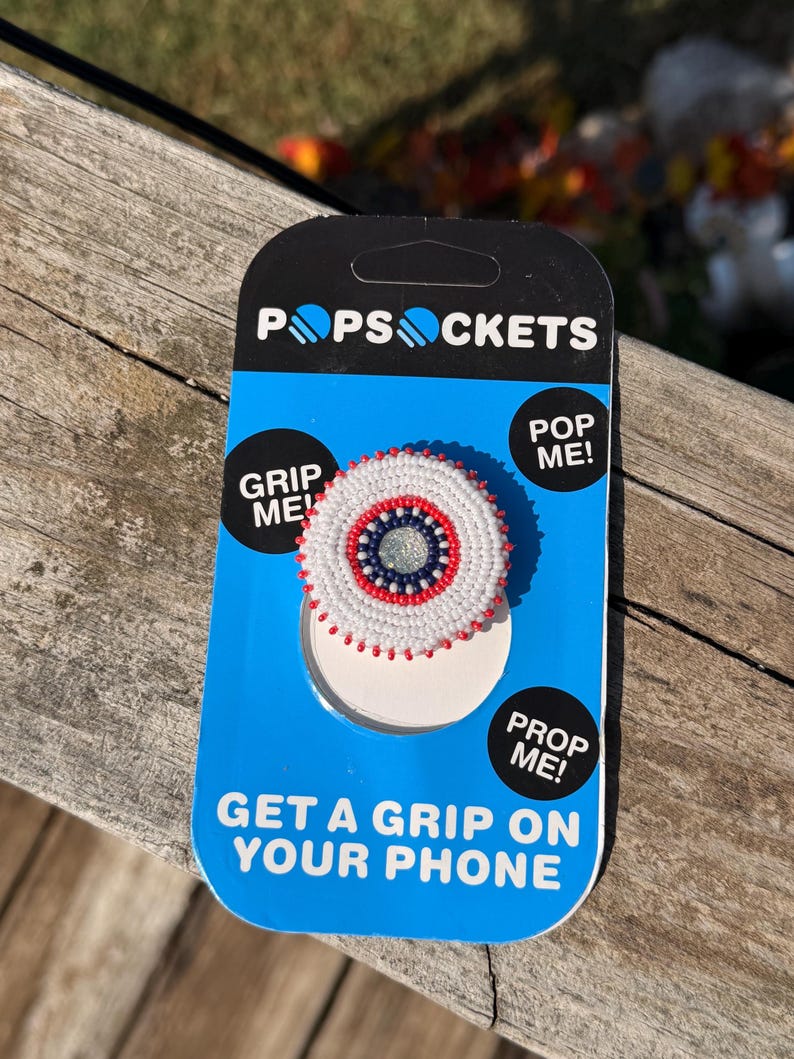 May include: A circular phone grip on a blue and black PopSockets display card. The grip features concentric rings of white, red, and dark blue seed beads surrounding a central silver glitter disc. The card includes the text PopSockets, Grip Me, Pop Me, Prop Me, and Get A Grip On Your Phone.