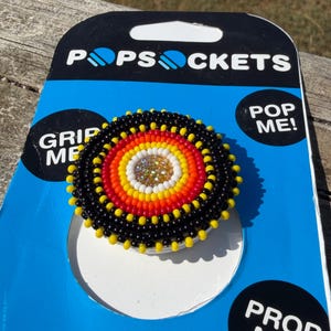 May include: A circular phone grip accessory featuring a beaded design with concentric rings of black, yellow, orange, and white beads surrounding a central cluster of clear crystals. The accessory is mounted on blue and black PopSockets packaging.