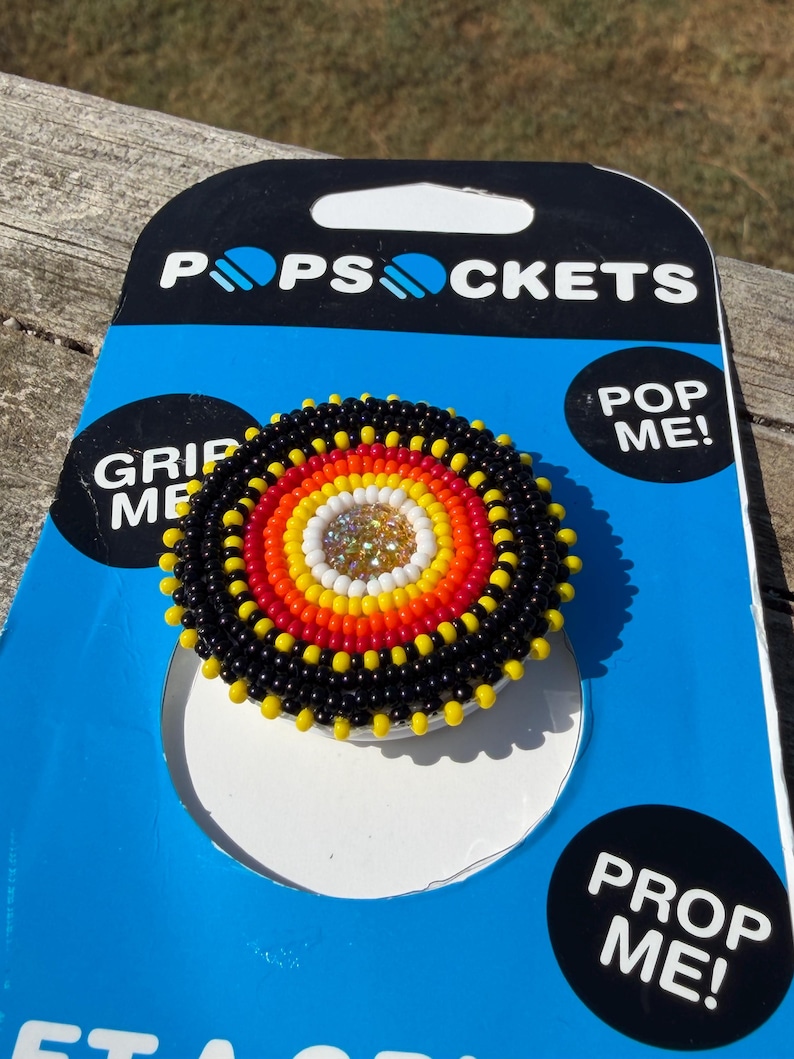 May include: A circular phone grip accessory featuring a beaded design with concentric rings of black, yellow, orange, and white beads surrounding a central cluster of clear crystals. The accessory is mounted on blue and black PopSockets packaging.