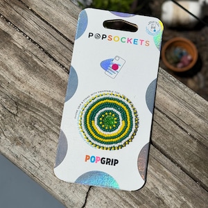 May include: A PopSockets PopGrip phone stand in its original white packaging. The circular grip features a concentric beaded pattern in alternating rings of green, yellow, and white, with a solid green center. The packaging includes the text PopSockets.