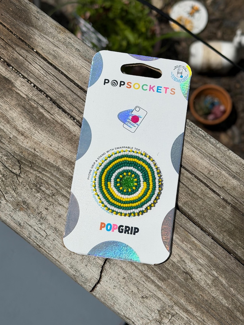 May include: A PopSockets PopGrip phone stand in its original white packaging. The circular grip features a concentric beaded pattern in alternating rings of green, yellow, and white, with a solid green center. The packaging includes the text PopSockets.
