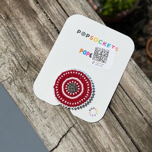 May include: A circular phone grip featuring a beaded design with concentric rings of red and white seed beads surrounding a dark gray center. The grip is attached to a white branded Popsockets display card with a QR code and colorful text.