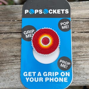 May include: A circular phone grip featuring a beaded design with concentric rings of red, orange, yellow, and white, centered with a red bead. It is mounted on a blue and black PopSockets brand display card with the text PopSockets, Grip Me, Pop Me, Prop Me, and Get A Grip On Your Phone.