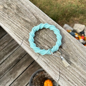 May include: A light blue macrame keychain wristlet with a silver-tone ring and a cream-colored tassel. The wristlet is made of twisted cotton cord. A small orange pumpkin sits in a pot below.
