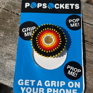 May include: A circular phone grip featuring concentric rings of black, red, orange, and white seed beads surrounding a central gold glitter accent. The accessory is mounted on a blue and black PopSockets retail card with the text PopSockets, Grip Me, Pop Me, Prop Me, and Get A Grip On Your Phone.
