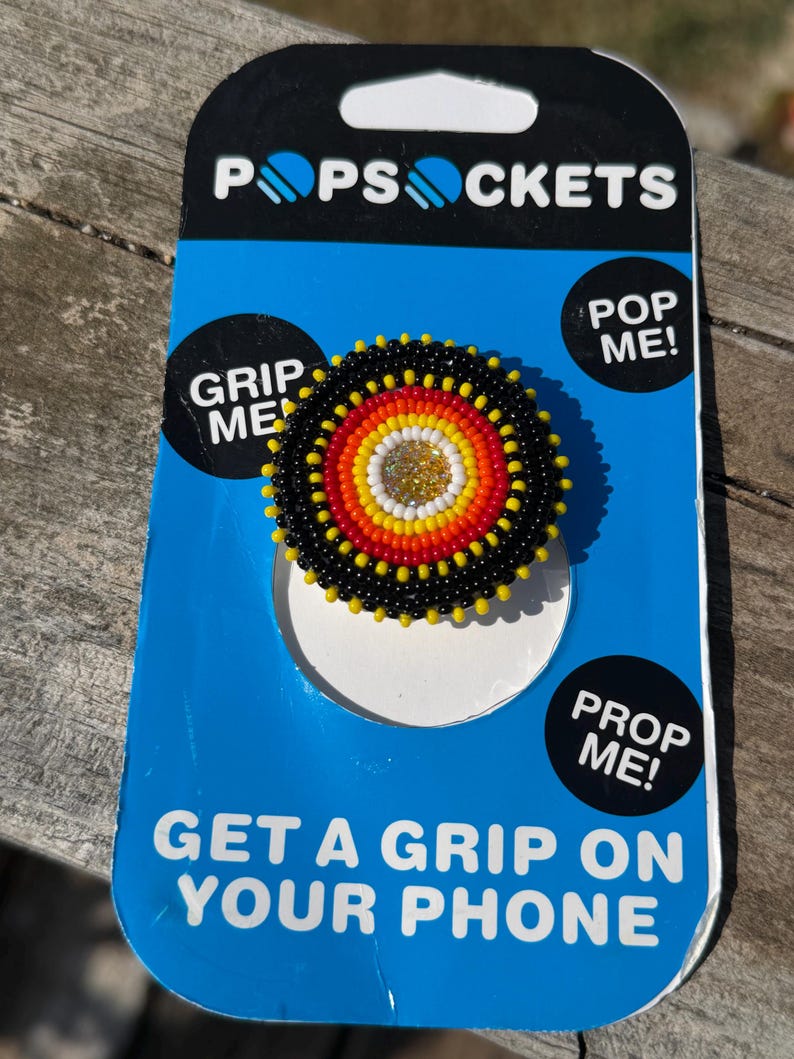 May include: A circular phone grip featuring concentric rings of black, red, orange, and white seed beads surrounding a central gold glitter accent. The accessory is mounted on a blue and black PopSockets retail card with the text PopSockets, Grip Me, Pop Me, Prop Me, and Get A Grip On Your Phone.