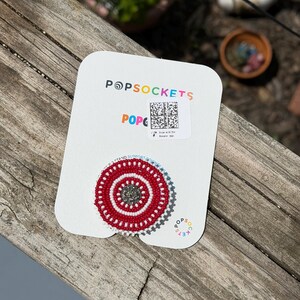 May include: A circular phone grip on a white display card with the text POPSOCKETS. The grip features a beaded design with a central dark gray circle surrounded by concentric rings of white and red beads. A QR code is printed on the card above.