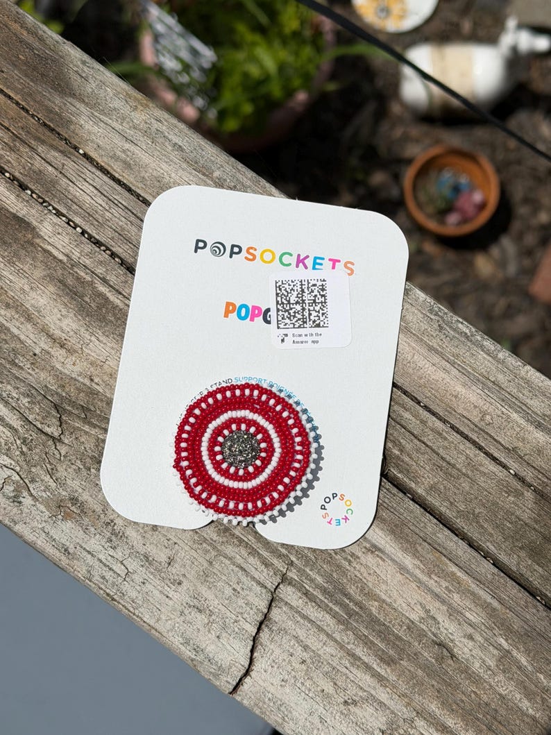 May include: A circular phone grip on a white display card with the text POPSOCKETS. The grip features a beaded design with a central dark gray circle surrounded by concentric rings of white and red beads. A QR code is printed on the card above.