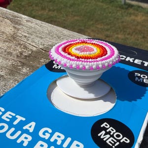 May include: A circular phone grip featuring a beaded top with concentric rings of pink, white, orange, and yellow beads surrounding a clear center. The white plastic base is mounted on blue packaging with the text GET A GRIP ON YOUR PHONE and PROP ME!