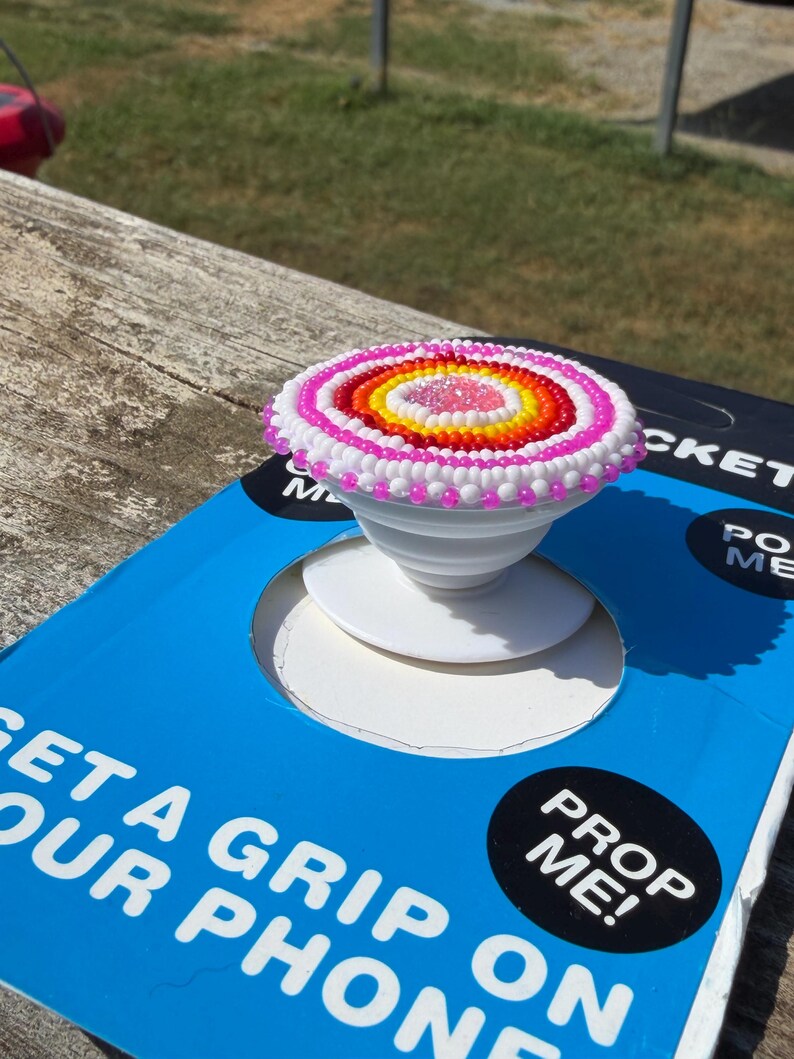 May include: A circular phone grip featuring a beaded top with concentric rings of pink, white, orange, and yellow beads surrounding a clear center. The white plastic base is mounted on blue packaging with the text GET A GRIP ON YOUR PHONE and PROP ME!