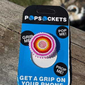 May include: A circular phone grip featuring concentric rings of seed beads in white, pink, orange, and yellow, with a glittery pink center. The accessory is mounted on a blue and black PopSockets branded retail card with the text PopSockets, Grip Me, Pop Me, Prop Me, and Get A Grip On Your Phone.
