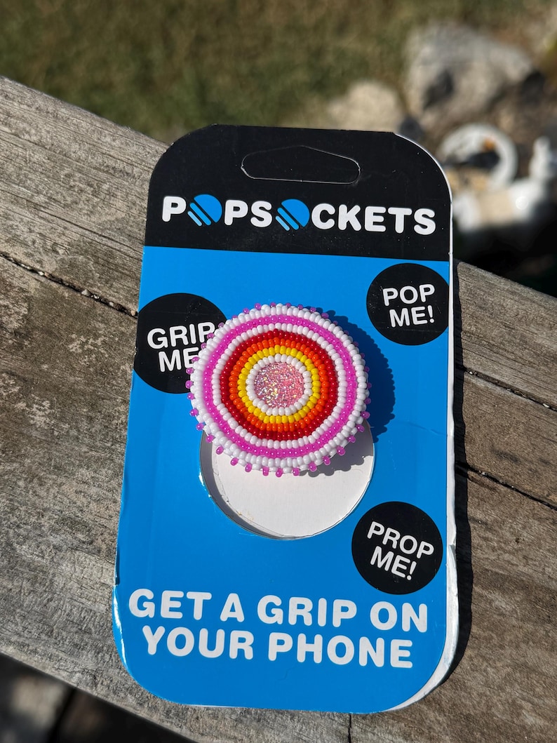 May include: A circular phone grip featuring concentric rings of seed beads in white, pink, orange, and yellow, with a glittery pink center. The accessory is mounted on a blue and black PopSockets branded retail card with the text PopSockets, Grip Me, Pop Me, Prop Me, and Get A Grip On Your Phone.