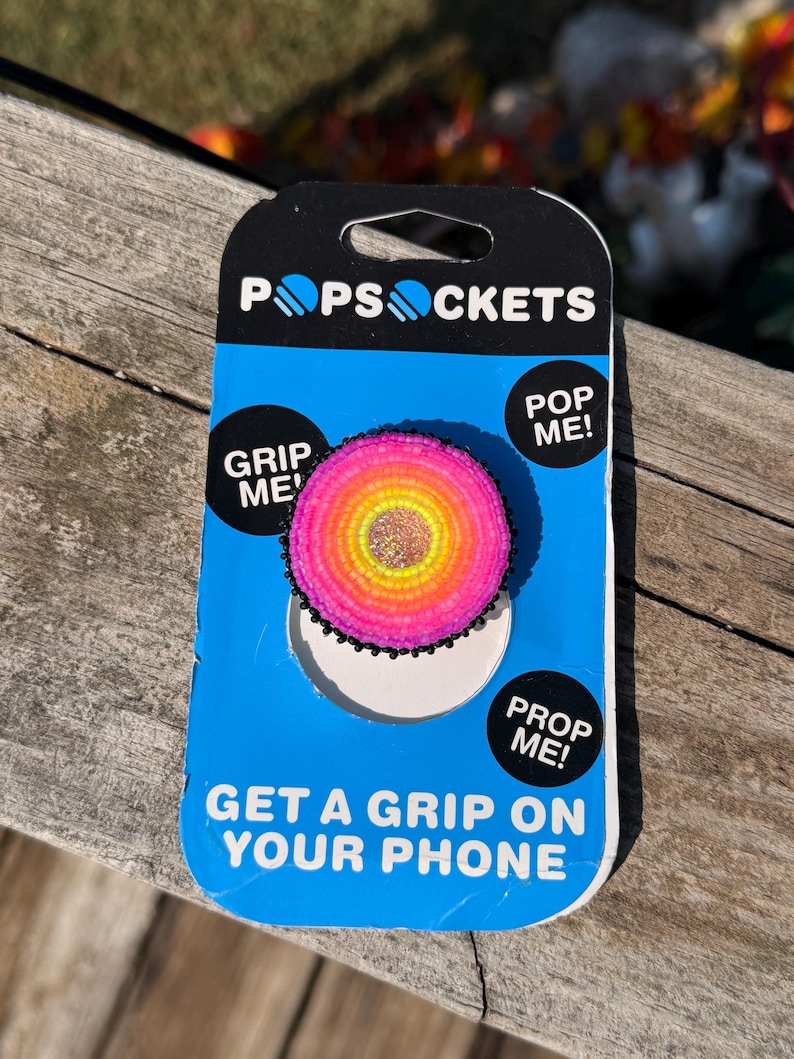 May include: A circular phone grip featuring a beaded design with concentric rings of pink, orange, and yellow, centered with a glittery gold disc. The grip is mounted on a blue and black PopSockets retail card with the text PopSockets, Grip Me, Pop Me, Prop Me, and Get a Grip on Your Phone.