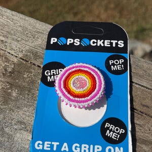 May include: A circular phone grip featuring concentric rings of small beads in pink, orange, yellow, and white, with a glittery pink center. The grip is attached to a blue and black PopSockets branded display card with the text PopSockets, Pop Me, Grip Me, Prop Me, and Get a Grip on Your Phone.