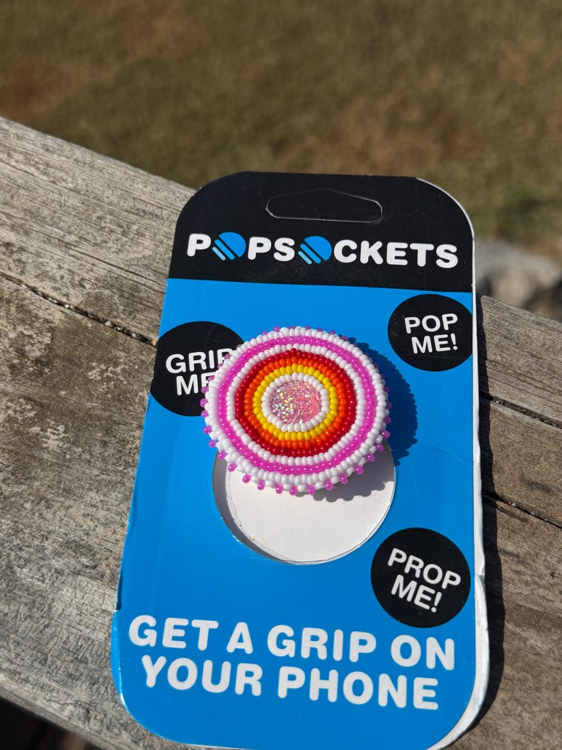 May include: A circular phone grip featuring concentric rings of small beads in pink, orange, yellow, and white, with a glittery pink center. The grip is attached to a blue and black PopSockets branded display card with the text PopSockets, Pop Me, Grip Me, Prop Me, and Get a Grip on Your Phone.