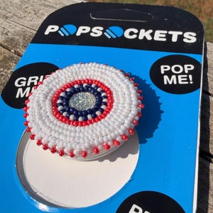 May include: A circular phone grip featuring intricate beadwork in concentric rings of white, red, and dark blue, centred with a clear faceted crystal. The grip is attached to its original blue and black PopSockets retail packaging card.
