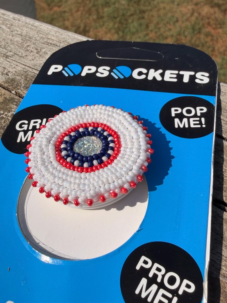 May include: A circular phone grip featuring intricate beadwork in concentric rings of white, red, and dark blue, centred with a clear faceted crystal. The grip is attached to its original blue and black PopSockets retail packaging card.