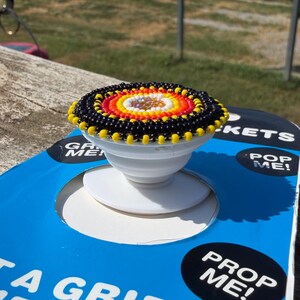 May include: A circular phone grip featuring a beaded top with concentric rings of black, yellow, orange, red, and white beads surrounding a central iridescent crystal. The base is a white collapsible plastic mount attached to blue packaging.