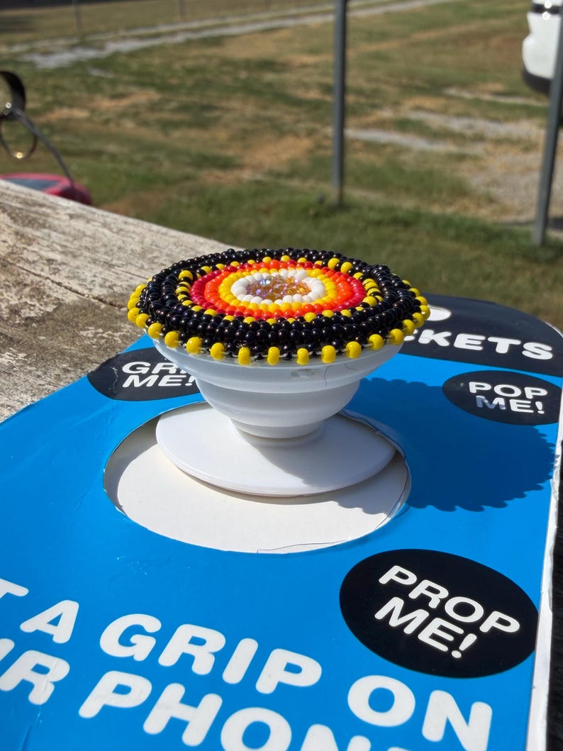 May include: A circular phone grip featuring a beaded top with concentric rings of black, yellow, orange, red, and white beads surrounding a central iridescent crystal. The base is a white collapsible plastic mount attached to blue packaging.