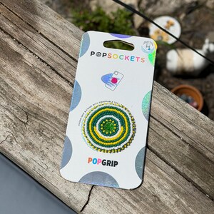 May include: A circular phone grip and stand on a white retail card with the text POPSOCKETS and POPGRIP. The accessory features a concentric beaded pattern in bright green, yellow, and white, with a small, solid green center circle.