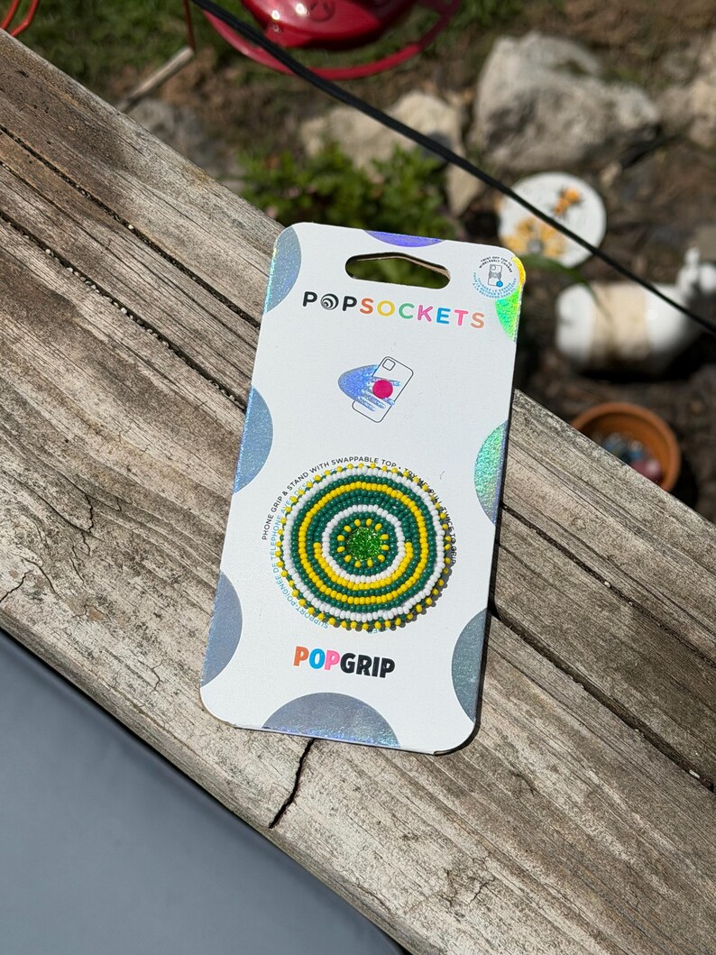 May include: A circular phone grip and stand on a white retail card with the text POPSOCKETS and POPGRIP. The accessory features a concentric beaded pattern in bright green, yellow, and white, with a small, solid green center circle.