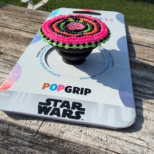 May include: A black phone grip featuring a circular top decorated with concentric rings of pink, black, and lime green beads. The phone grip is attached to a white retail display card with the text POPGRIP and STAR WARS printed in black.