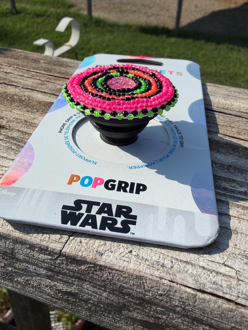 May include: A black phone grip featuring a circular top decorated with concentric rings of pink, black, and lime green beads. The phone grip is attached to a white retail display card with the text POPGRIP and STAR WARS printed in black.