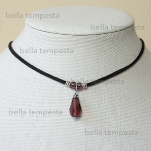 May include: A black choker necklace with a small, teardrop-shaped pendant made of purple glass. The pendant is attached to the necklace with silver beads.