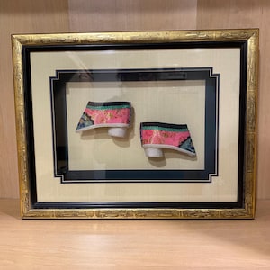 May include: Framed display of two antique Chinese lotus shoes. The shoes are pink with floral embroidery, black trim, and white soles. The frame is gold with a black and cream matting.