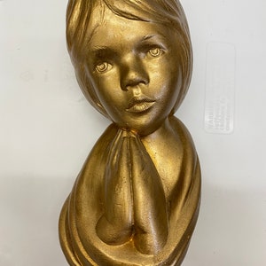 May include: A gold-colored ceramic sculpture of a child with their hands clasped in prayer. The sculpture is detailed with realistic features and a flowing robe.